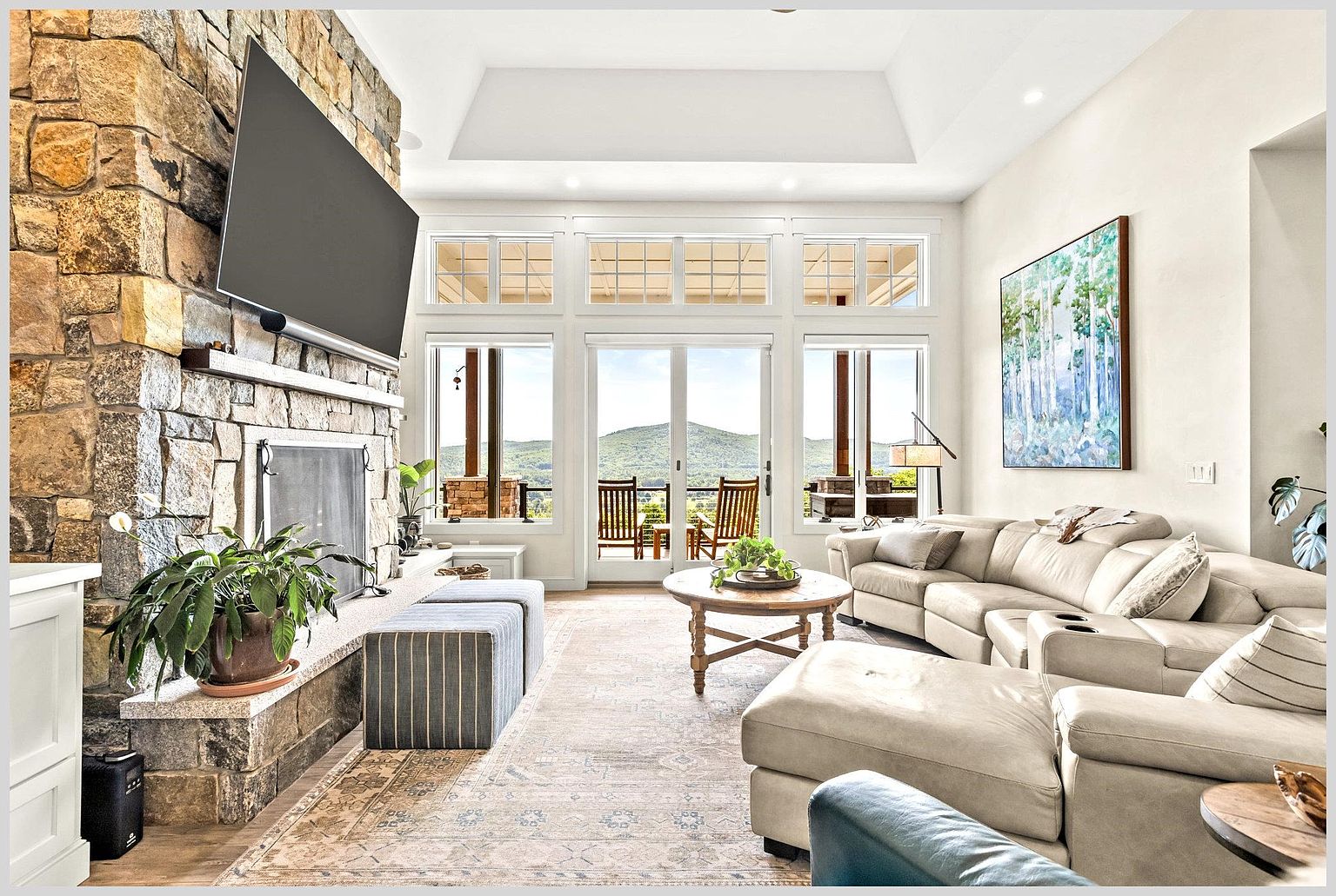This spacious living room features a prominent stone-clad chimney breast and a large television mounted above the mantel. A neutral-toned sectional sofa and a round wooden coffee table create a comfortable seating area, complemented by a large patterned area rug. Tall windows and glass doors provide abundant natural light and frame a scenic view of the distant mountains.