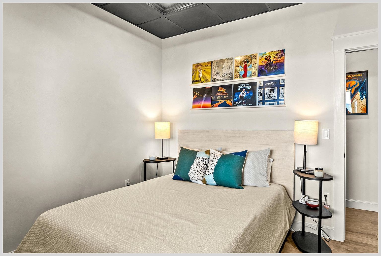 This cozy guest bedroom features a light wood-toned headboard centered against a neutral wall, accented by a collection of framed album covers displayed on a wall-mounted ledge. Two small, tiered side tables with matching lamps flank the bed, which is dressed in a neutral-toned comforter and decorative teal and patterned pillows. The room is finished with light wood flooring and a dark, coffered-style ceiling, creating a modern and personalized aesthetic.