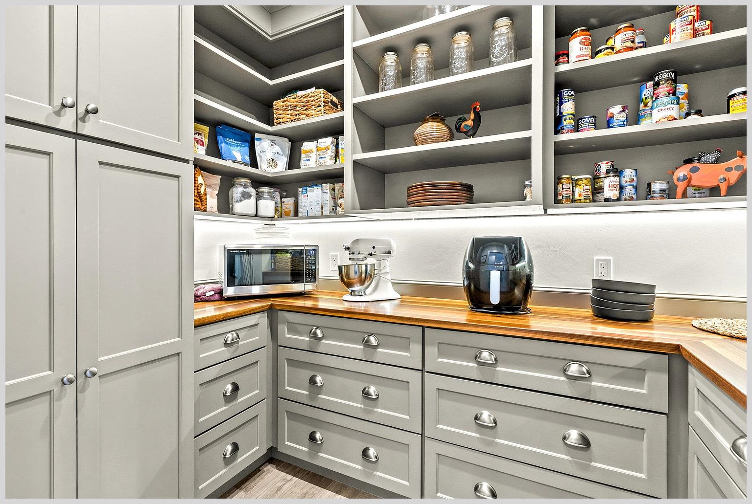 This organized walk-in pantry features light gray cabinetry with brushed metal hardware and a warm-toned wood countertop. The space is equipped with a microwave, a stand mixer, and an air fryer, while open shelving provides ample storage for canned goods and jars. The clean, functional design creates a bright and efficient storage area perfect for a modern home.