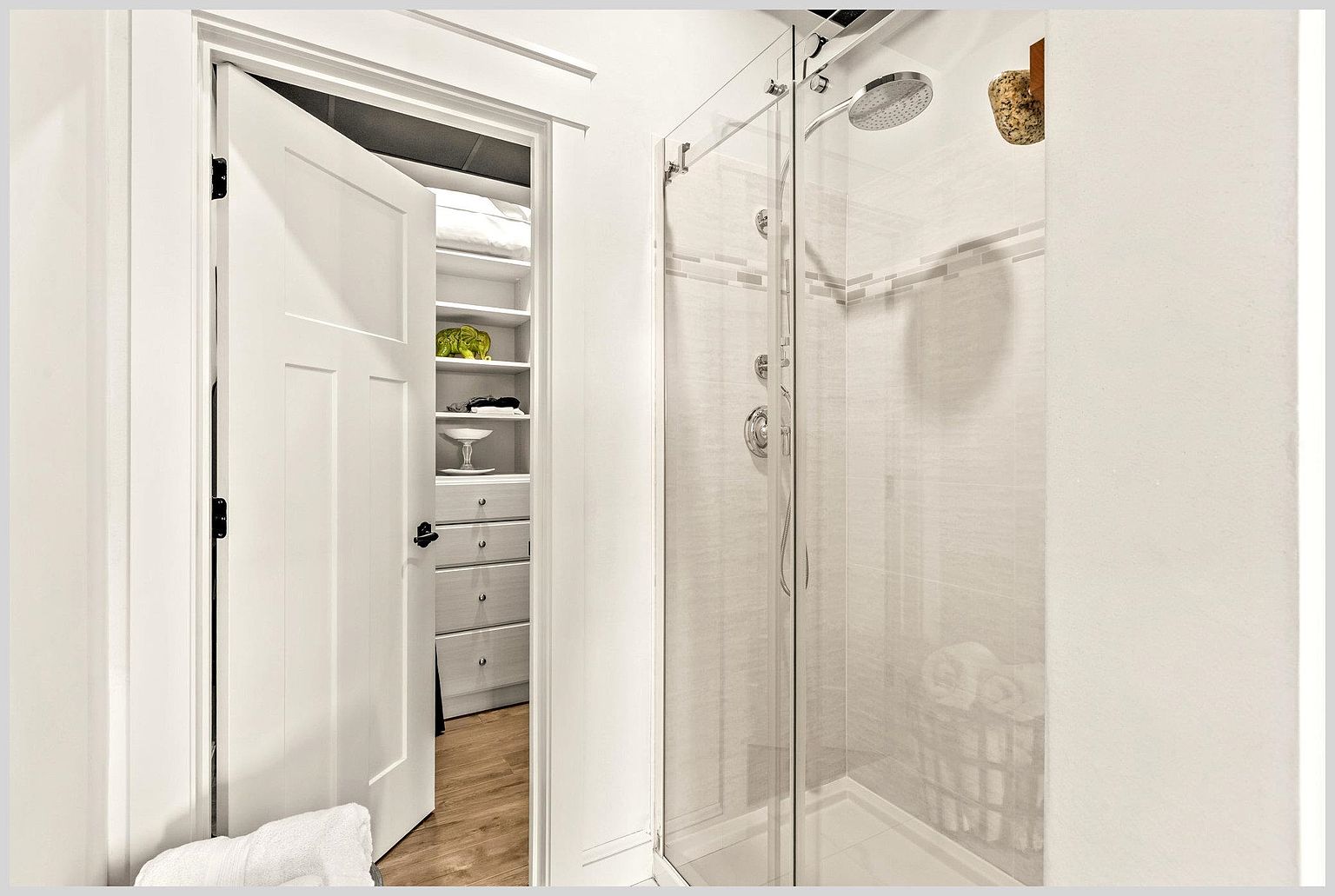This image captures a bright, modern bathroom featuring a glass-enclosed walk-in shower with a rainfall showerhead and wall-mounted fixtures. To the left, an open white door reveals a walk-in closet with organized shelving and drawers, creating a seamless transition between the two spaces. The neutral color palette and clean lines provide a contemporary and functional aesthetic.