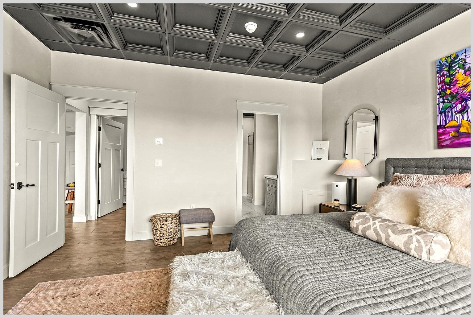This primary bedroom features a striking coffered ceiling painted in a dark, moody tone that contrasts with the bright white walls. The room is furnished with a plush gray upholstered bed, a textured rug, and a small stool, creating a cozy and inviting atmosphere. A doorway leads to an adjacent bathroom, while a second door opens to a hallway, highlighting the home's open and connected layout.