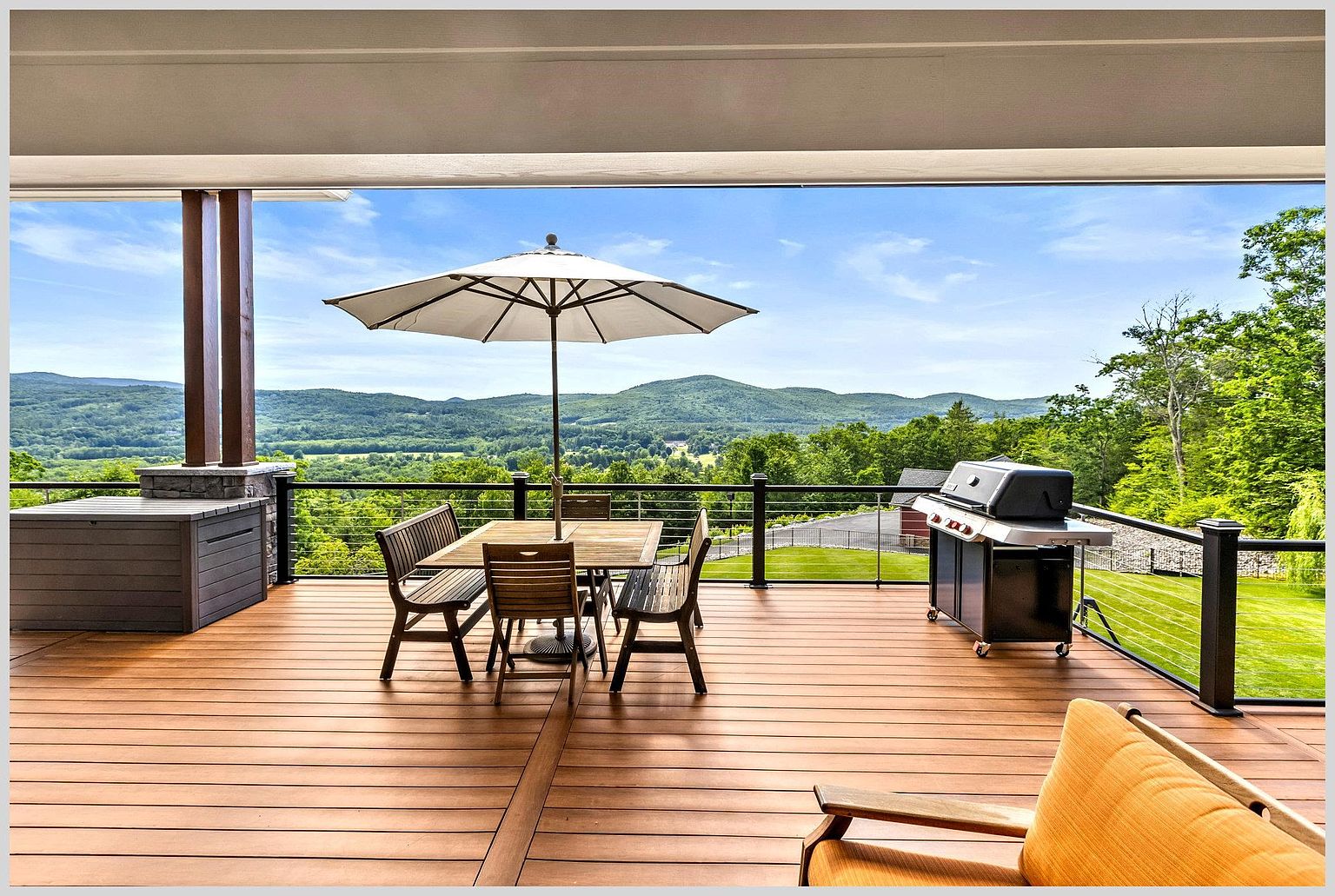 This expansive, elevated deck offers a panoramic view of rolling green hills and a lush landscape under a bright, clear sky. The space is furnished with a wooden dining set shaded by a large umbrella and a stainless steel grill, making it an ideal setting for outdoor entertaining. The deck features modern cable railings that maintain an unobstructed view, creating a seamless transition between the home and the natural scenery.
