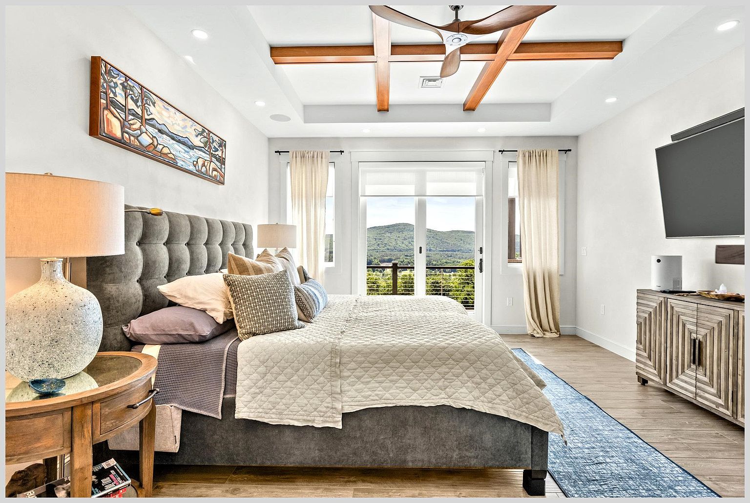 This primary bedroom features a sophisticated design with a tufted gray upholstered headboard and a neutral-toned quilt. The room is highlighted by a coffered ceiling with exposed wood beams and a central ceiling fan, while large glass doors provide a scenic view of the surrounding mountains. A patterned wooden dresser sits beneath a wall-mounted television, and a blue runner rug adds a touch of color to the light wood-toned flooring.