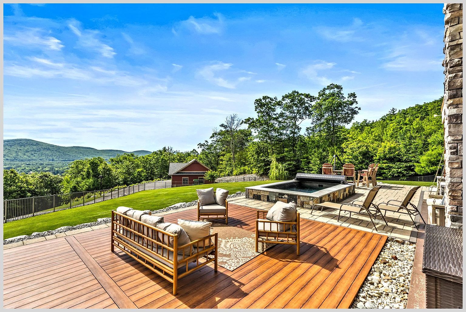 This expansive outdoor living space features a multi-level deck with warm-toned wood decking and a stone-clad hot tub area. The patio is furnished with comfortable lounge seating and sun loungers, overlooking a manicured lawn and a scenic mountain landscape. The scene is captured from a slightly elevated perspective, emphasizing the seamless transition between the home's architecture and the natural surroundings.
