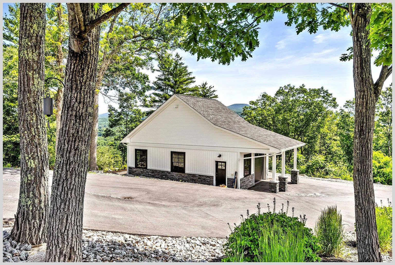 This image captures a detached garage building with a clean, modern farmhouse aesthetic, featuring white vertical siding and a dark stone base. The structure includes a covered side porch area with stone-clad pillars and a paved driveway that winds through a wooded, elevated landscape. The perspective is a wide-angle exterior shot that highlights the building's integration into the natural, scenic environment.