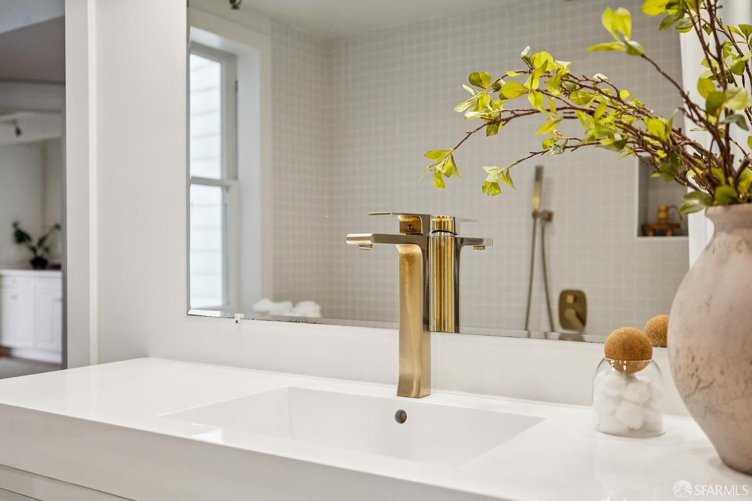 This image showcases a modern bathroom featuring a sleek, white countertop with an integrated sink and a stylish gold faucet. A large mirror reflects the bathroom's design, including a tiled wall and a glimpse of a shower area. The presence of decorative elements like a vase with greenery and a jar of cotton balls adds a touch of elegance and sophistication.