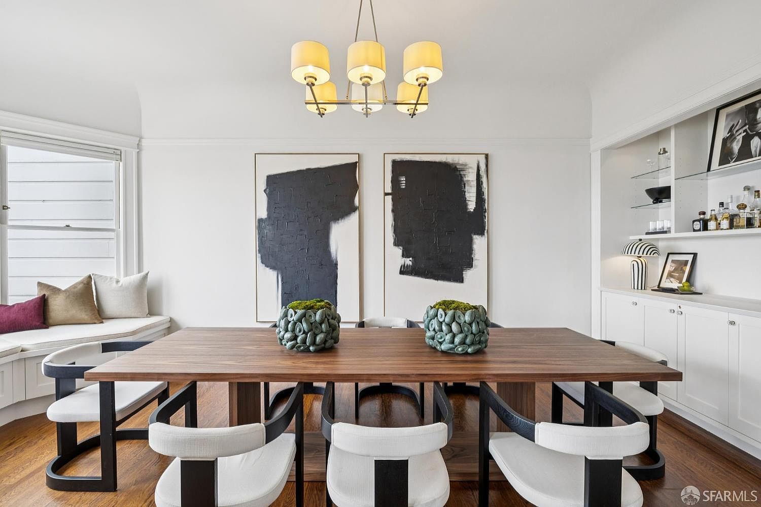 This is an interior shot of a dining room featuring a long wooden table with modern chairs. Two large abstract paintings hang on the wall, complemented by decorative vases on the table. A built-in window seat and shelving unit add to the room's charm and functionality, creating an inviting space for dining and entertaining.