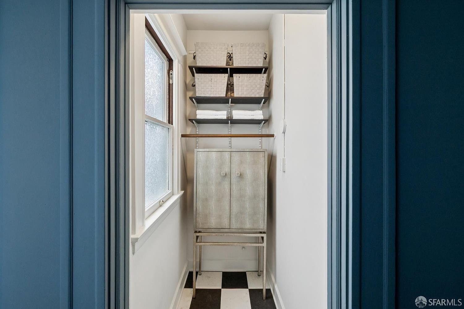 The image showcases a well-organized walk-in closet. It features a cabinet with a silver finish, shelves holding woven baskets and neatly folded towels, and a black and white checkered floor. A window provides natural light, enhancing the clean and functional design of the space.