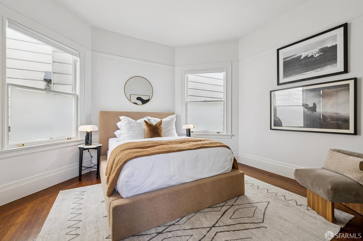 This is a bright and inviting primary bedroom featuring a neutral color palette. The bed has a tan upholstered frame and is adorned with white linens, a brown throw blanket, and decorative pillows. Natural light streams in through the windows, and artwork hangs on the walls, adding a touch of sophistication.