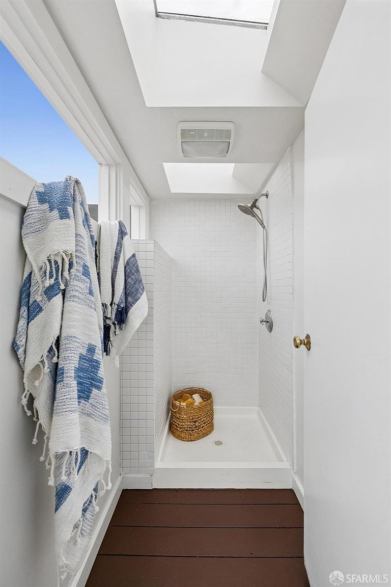 This is a bright and compact bathroom featuring a walk-in shower with white subway tile and a white shower base. A window provides natural light, complemented by a skylight above the shower. A woven basket sits on the shower floor, and a blue and white patterned towel hangs near the window, adding a touch of color.