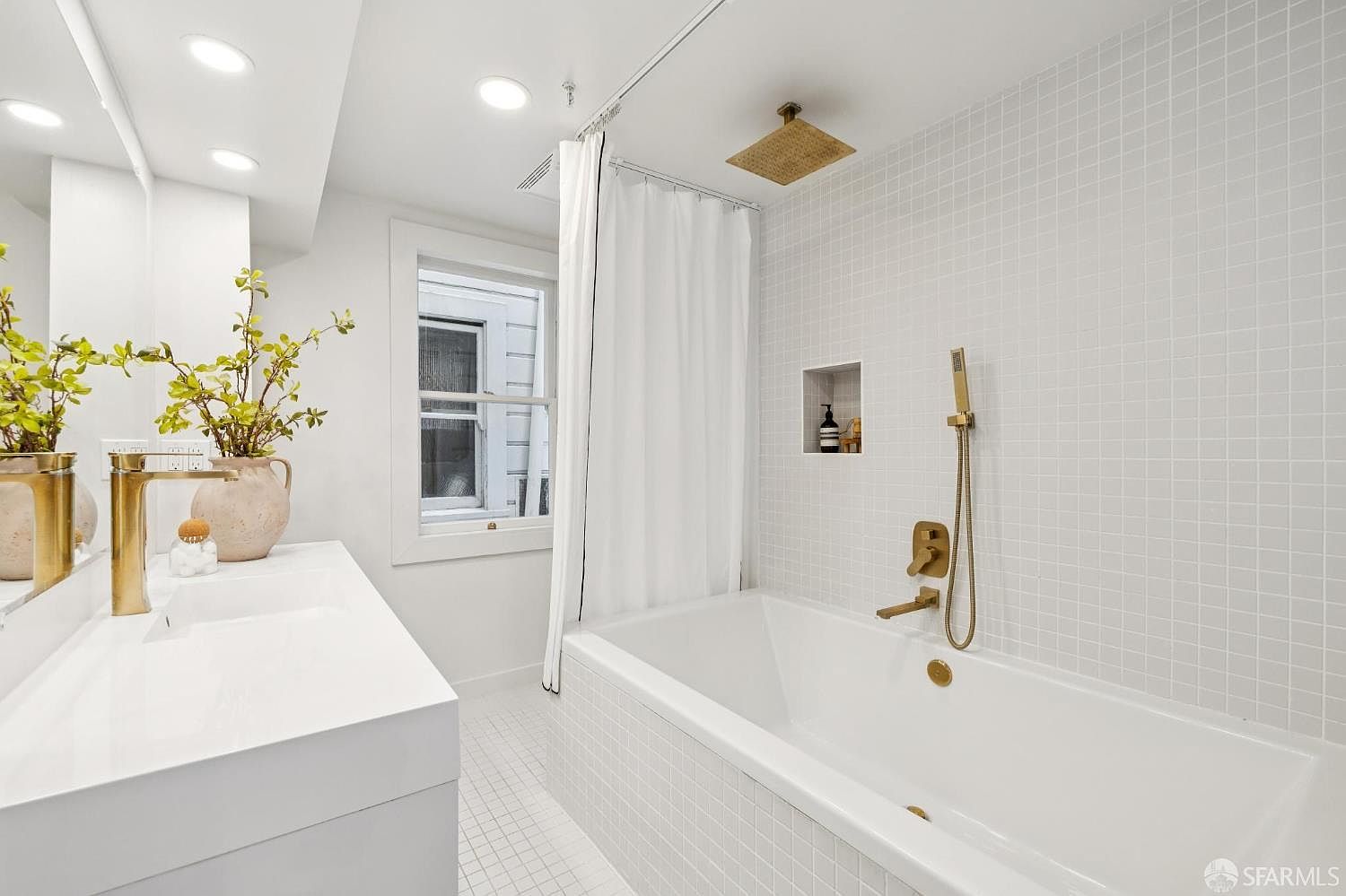 This is a bright and modern bathroom featuring white square tiles on the walls and floor. A white bathtub is positioned next to a window with a white curtain, and gold fixtures add a touch of luxury. The vanity has a white countertop with a gold faucet and decorative plants.