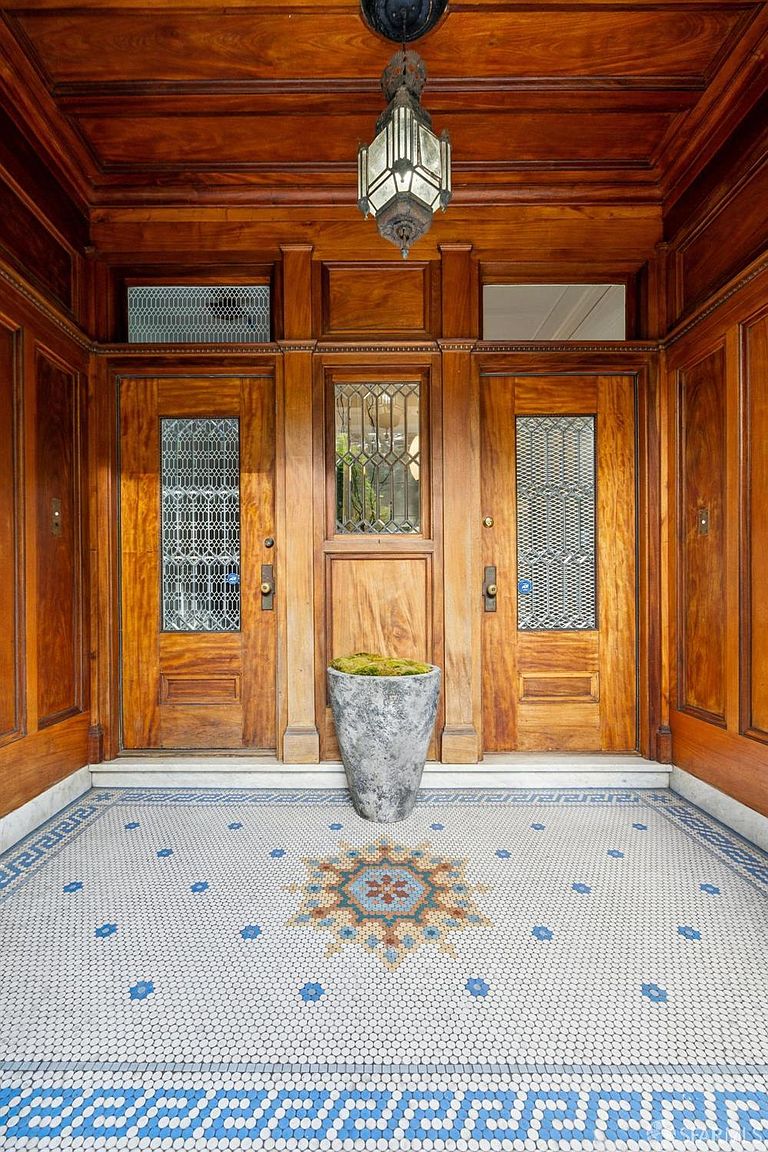 This image showcases a grand entryway with rich, dark wood paneling and doors, creating a luxurious and inviting first impression. The intricate tile flooring features a decorative central medallion and a Greek key border, adding a touch of elegance. A large, textured planter sits in front of the center door, enhancing the visual appeal.