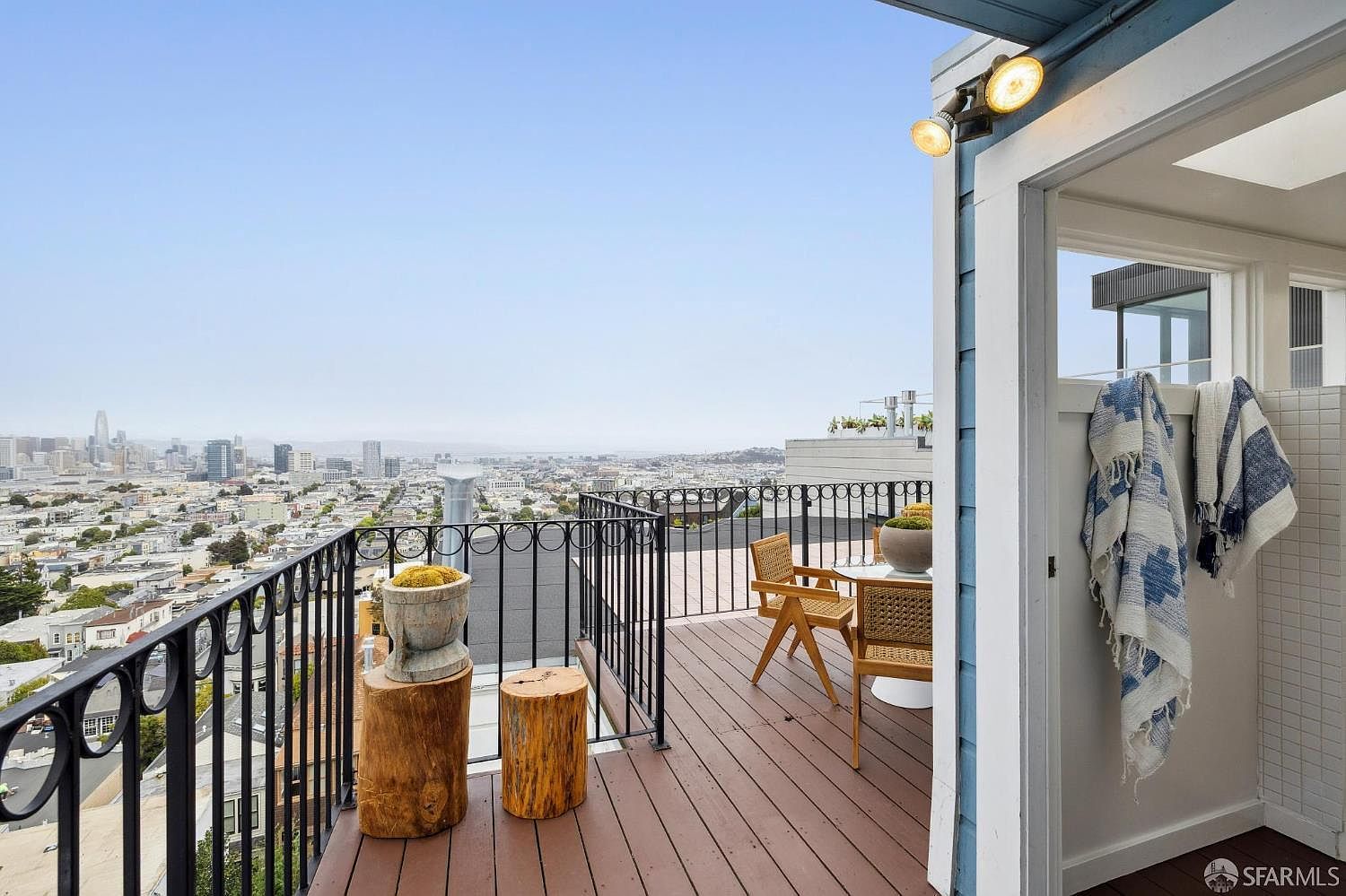 This image showcases a charming patio/deck/balcony with a stunning city view. The space features wooden decking, a black metal railing, and outdoor furniture including a small table and chairs. Adjacent to the deck is an outdoor shower area, adding a touch of luxury and convenience to the outdoor space.
