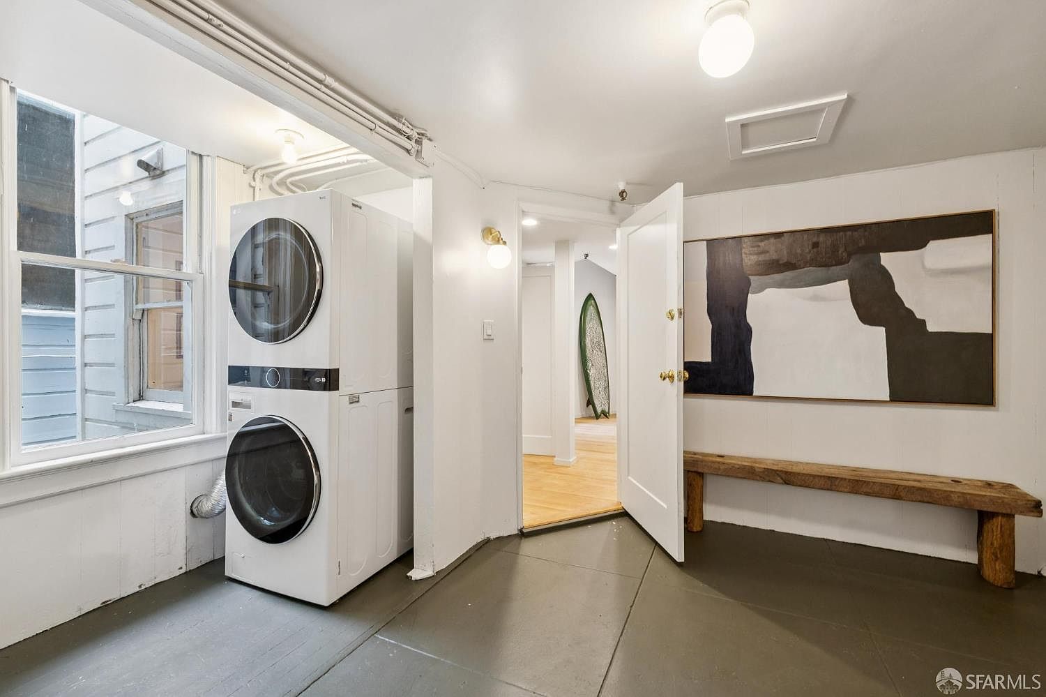 This is an interior shot of a laundry room featuring a stacked washer and dryer set next to a window. The room has a clean, minimalist aesthetic with white walls and trim, contrasted by a dark gray floor. An open doorway leads to another room, and a piece of abstract art hangs above a wooden bench, adding a touch of sophistication.
