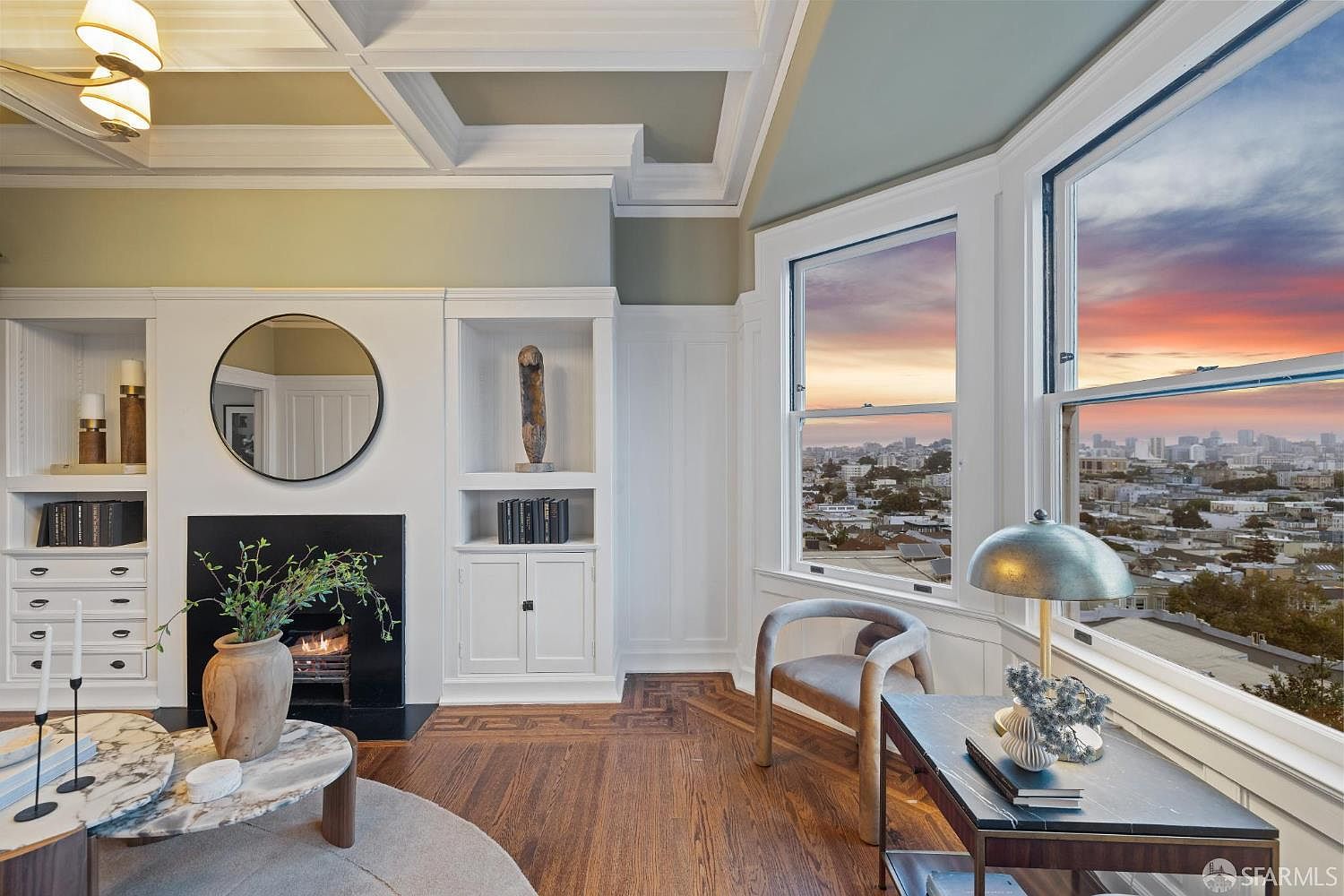 This is an interior shot of a living room featuring a fireplace with built-in shelving, a round mirror above the fireplace, and a bay window offering a city view at sunset. The room is decorated in a classic style with hardwood floors, a coffered ceiling, and neutral-toned furniture, creating a warm and inviting atmosphere.