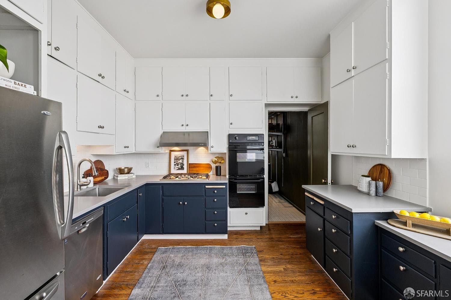 This is a well-lit kitchen featuring two-tone cabinetry, with white upper cabinets and navy blue lower cabinets. Stainless steel appliances, including a refrigerator and oven, are visible. The kitchen has hardwood floors and a gray rug, creating a warm and inviting atmosphere.