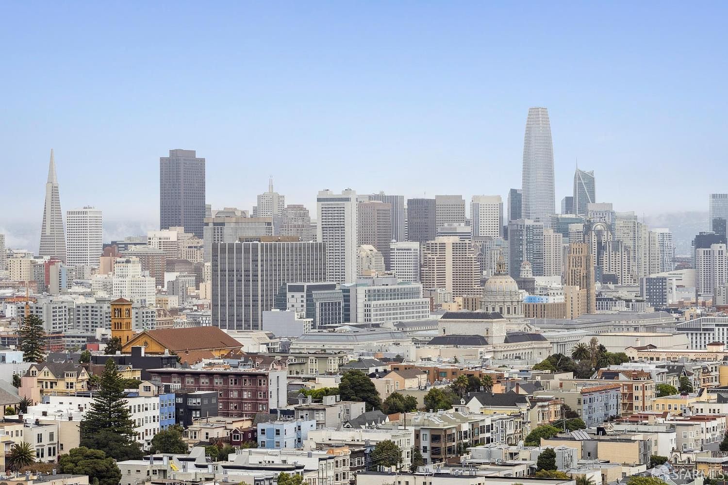 This aerial view showcases the San Francisco skyline, dominated by modern skyscrapers and iconic landmarks. The cityscape is set against a clear blue sky, with residential neighborhoods visible in the foreground, offering a sense of urban density and vibrant city living. The image provides a comprehensive overview of the city's architectural diversity and scale.