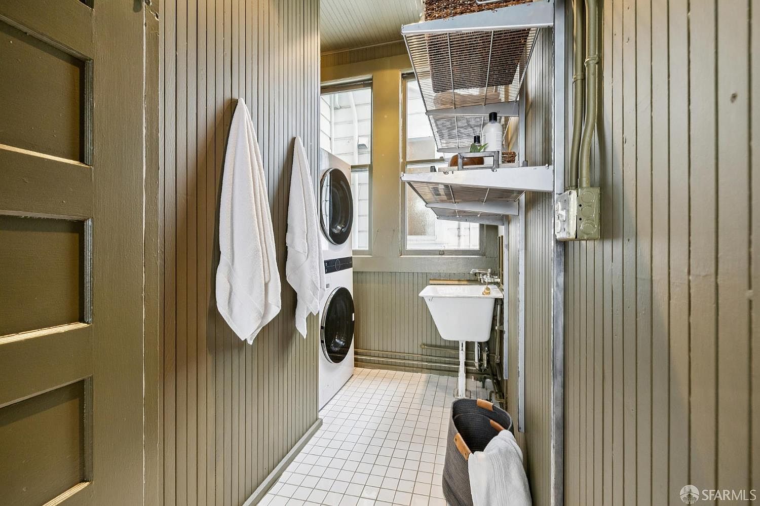 This is an interior shot of a laundry room featuring a stacked washer and dryer set against a vertically paneled wall with hanging white towels. A utility sink and wire shelving are visible on the right, and the room is illuminated by natural light filtering through a window. The floor is tiled in a white grid pattern, and the walls are painted in a muted green tone.