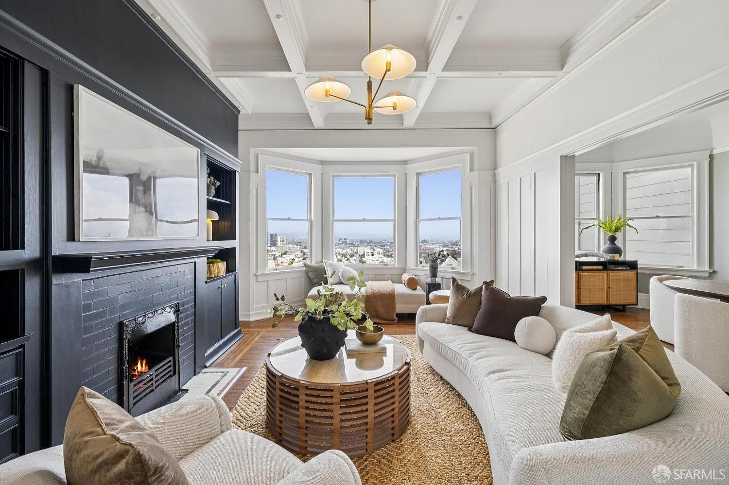 This is a beautifully staged living room featuring a black fireplace with a large art piece above it, a curved white sofa with decorative pillows, and a round coffee table on a jute rug. The room has a bright and airy feel, enhanced by the large bay windows offering city views and the coffered ceiling with a modern chandelier. The overall impression is one of sophisticated comfort and style.