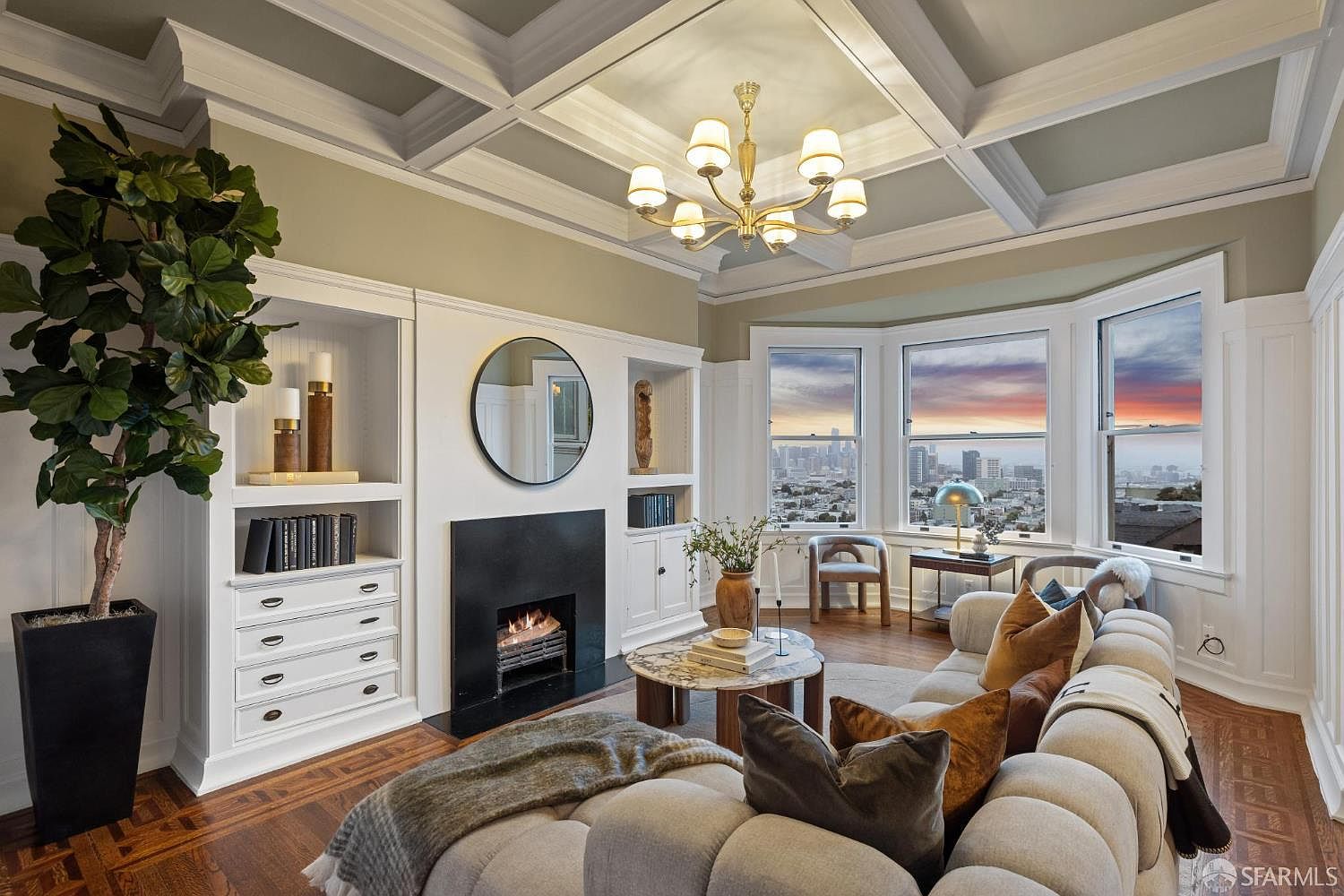 This is an interior shot of a living room featuring a fireplace, built-in shelving, and a bay window with a city view. The room is decorated in a classic style with a modern twist, featuring a round sofa and a coffered ceiling with a chandelier. The overall impression is one of elegance and comfort.