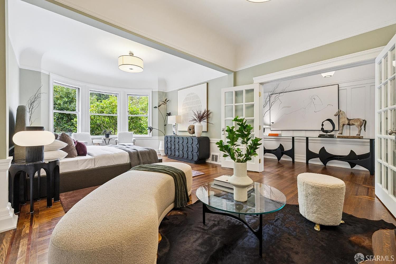 This is a stylish primary bedroom featuring a bed with neutral bedding, a curved bench, and a glass-topped coffee table on a dark rug. The room has hardwood floors, large windows providing natural light, and elegant decor including artwork and a modern dresser. The overall impression is luxurious and inviting.