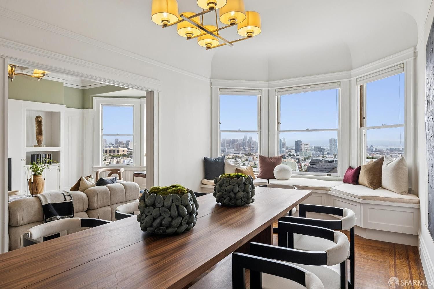 This is an interior shot of a dining room featuring a long wooden table with modern chairs. The room has a bay window with a cushioned seating area offering a city view. A modern chandelier hangs above the table, and decorative moss-filled stone bowls serve as centerpieces.