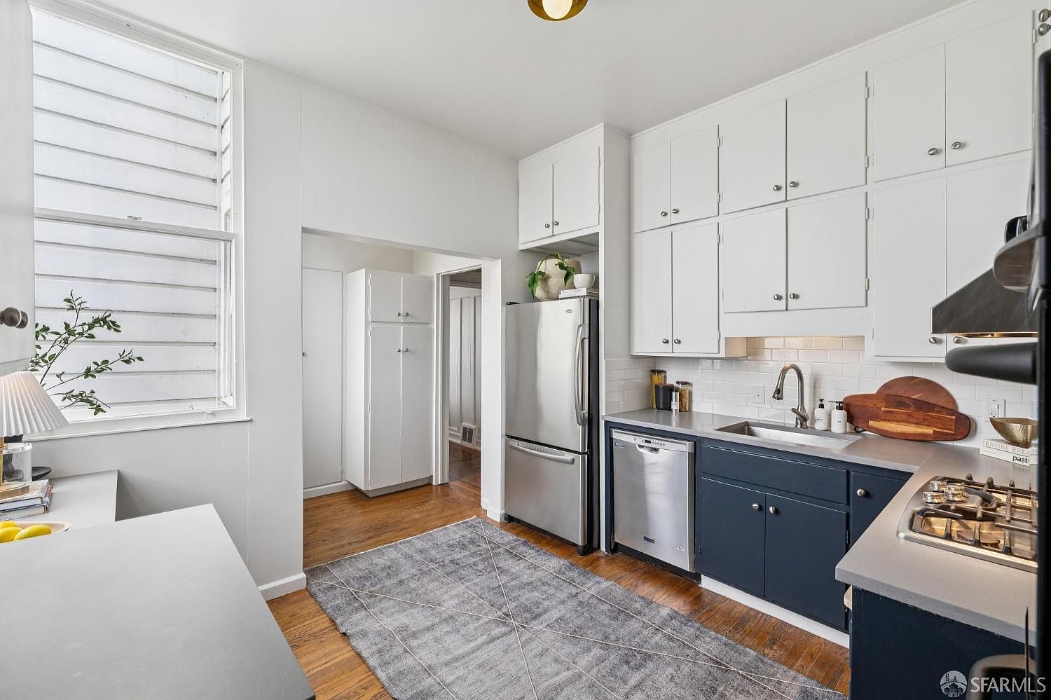 This is a well-lit kitchen featuring white upper cabinets and navy blue lower cabinets, complemented by stainless steel appliances. A gray patterned rug adds texture to the hardwood floors, and the overall design presents a clean and functional space. The perspective is from a medium distance, capturing the layout and key elements of the kitchen.