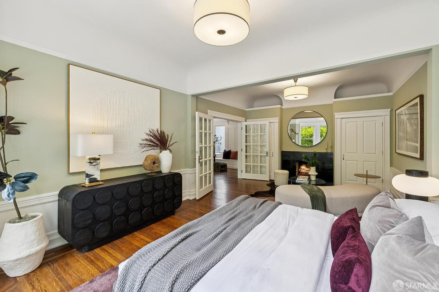 This is a luxurious primary bedroom featuring hardwood floors, a large bed with plush pillows and a gray throw, and a dark wood dresser with decorative items. A large white abstract art piece hangs above the dresser. The room opens into a cozy sitting area with a fireplace and a round mirror, creating a serene and inviting atmosphere.