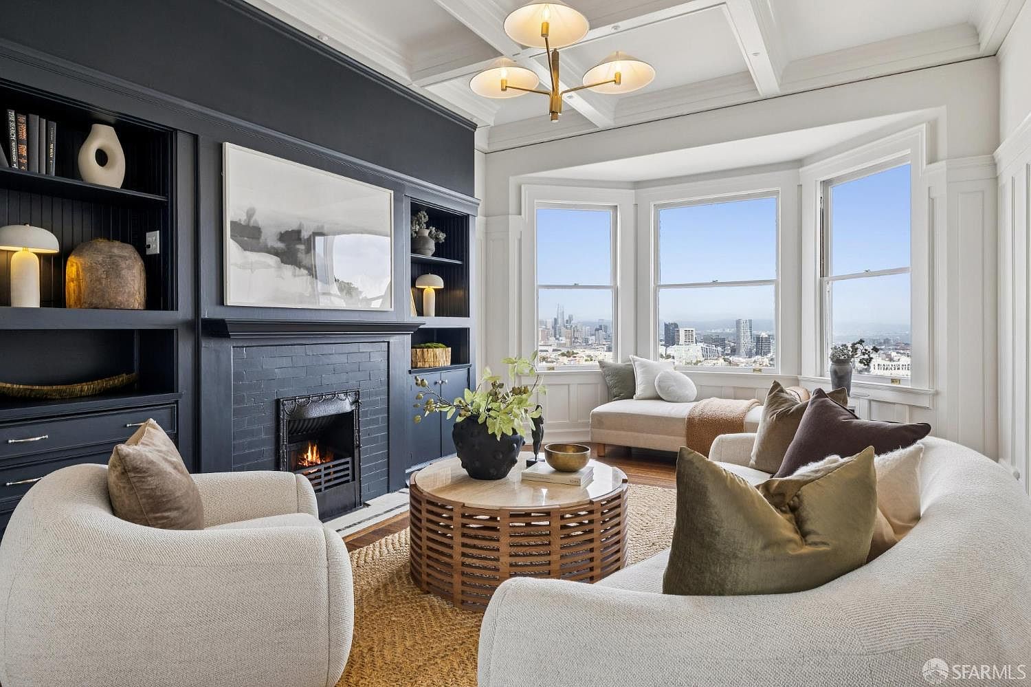 This is an interior shot of a living room featuring a dark-painted fireplace with built-in bookshelves on either side. The room is furnished with comfortable, light-colored seating arranged around a round, wooden coffee table. Large windows offer a stunning city view, and a modern chandelier hangs from the coffered ceiling, creating a sophisticated and inviting atmosphere.