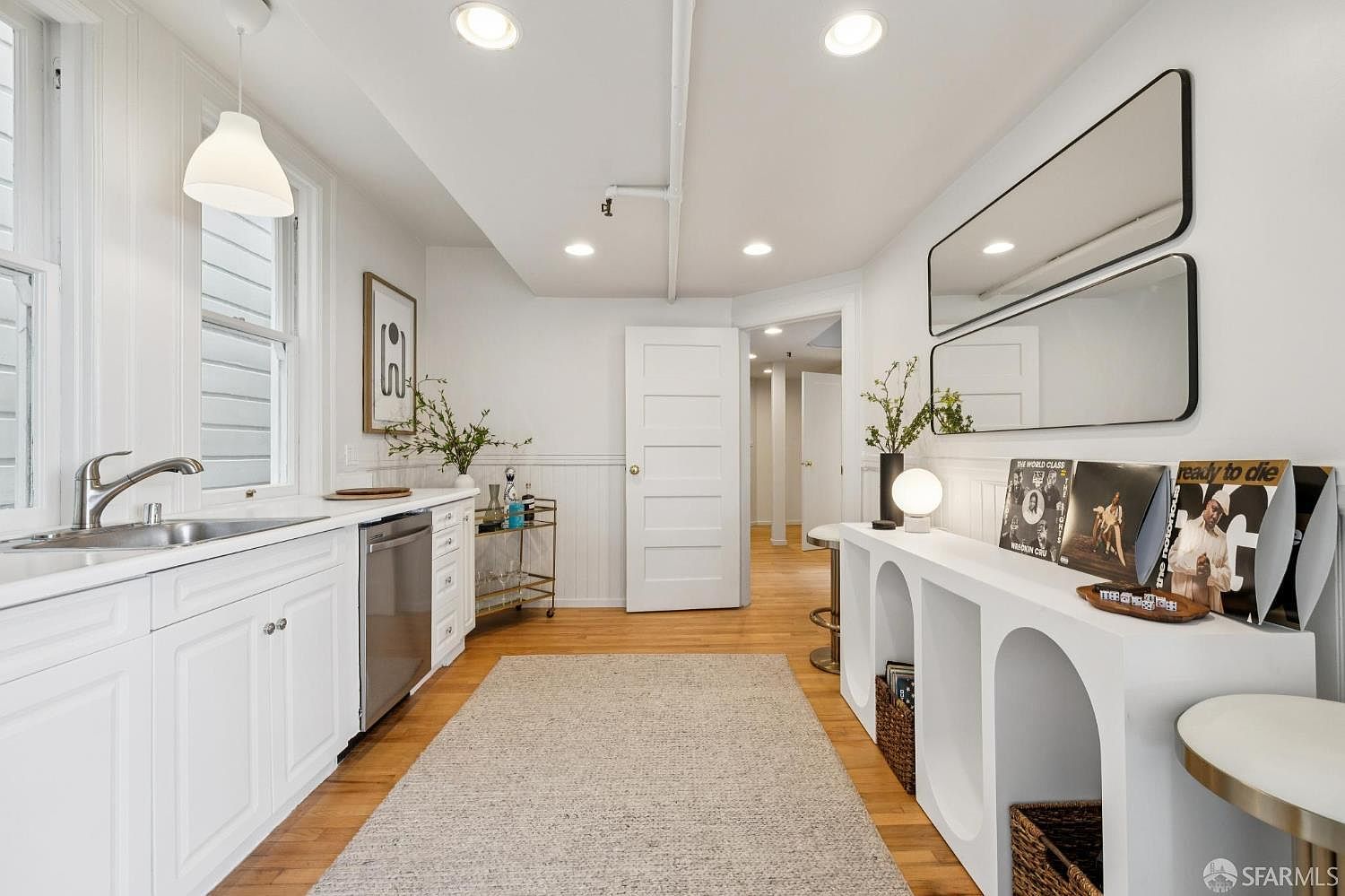 This is a bright and modern kitchen area featuring white cabinetry, stainless steel appliances, and hardwood floors. The space is well-lit with recessed lighting and a pendant light, and it includes a stylish bar cart and a unique display shelf with decorative items. The overall impression is clean, organized, and inviting.