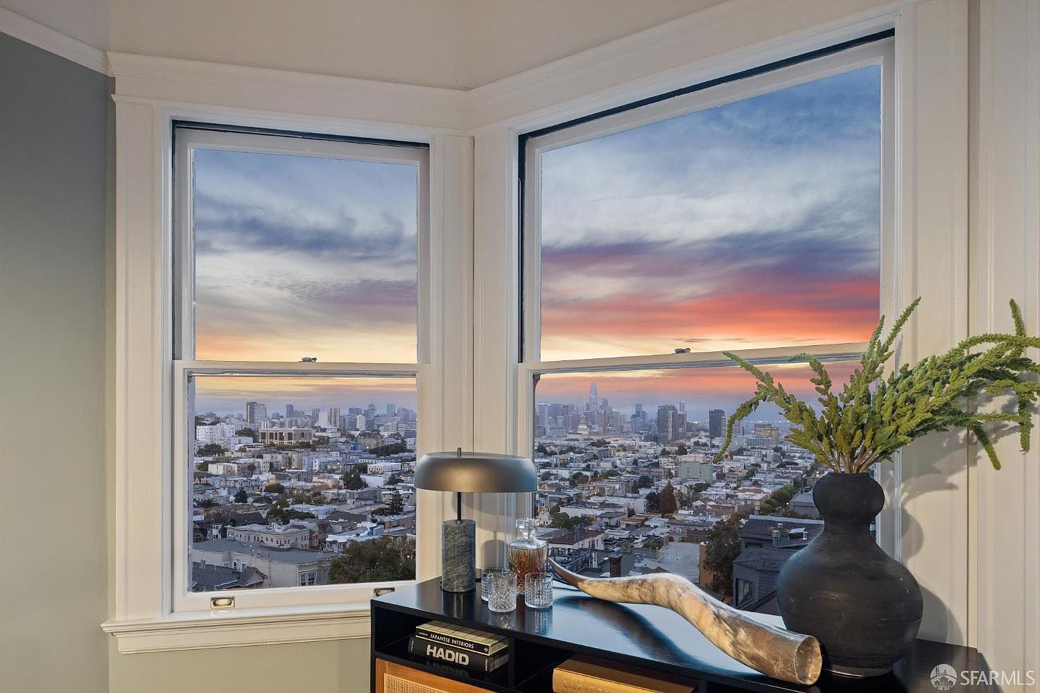 This image showcases a cozy interior space with a stunning city view through two large windows. A dark-colored cabinet displays decorative items such as a lamp, books, a decanter set, a horn, and a vase with greenery. The room exudes a sophisticated and comfortable ambiance, highlighting the property's prime location and stylish decor.
