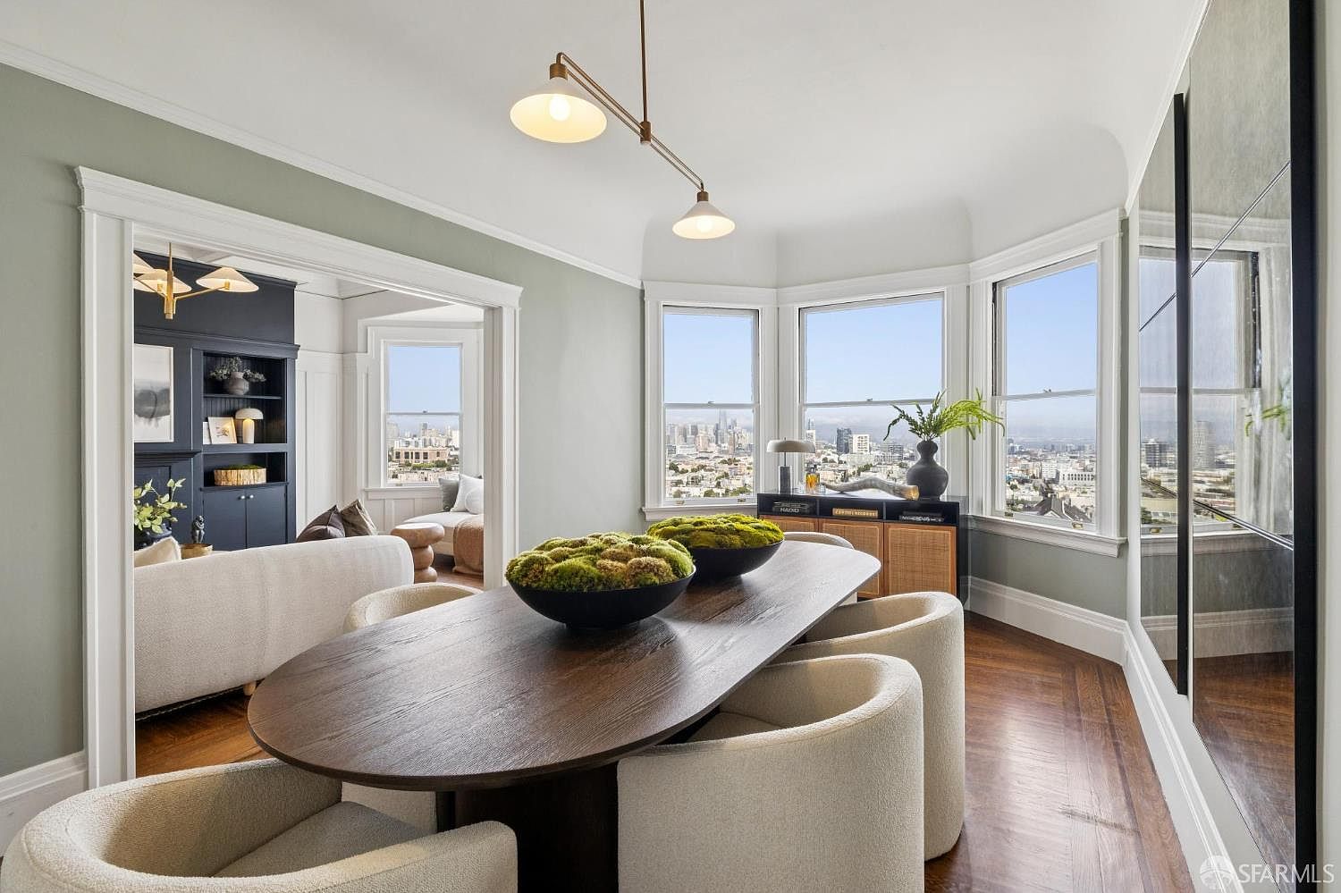 This is an interior shot of a dining room featuring a long, oval-shaped wooden table with modern, upholstered chairs. Two decorative moss bowls sit atop the table, and a contemporary light fixture hangs above. Large windows offer a stunning city view, enhancing the room's appeal and creating a bright, airy atmosphere.