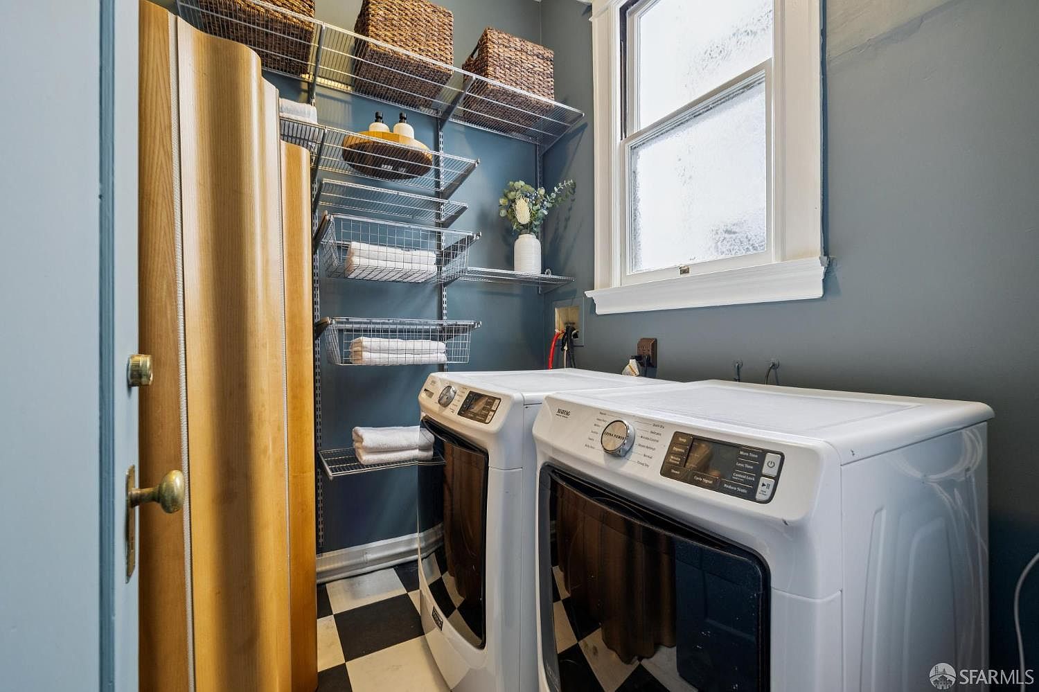 This is an interior shot of a laundry room featuring a black and white checkered floor, a modern white washer and dryer set, and wire shelving with baskets and towels. The walls are painted a muted blue-gray, and a window provides natural light. A wooden door frame is visible on the left, adding a touch of warmth to the space.