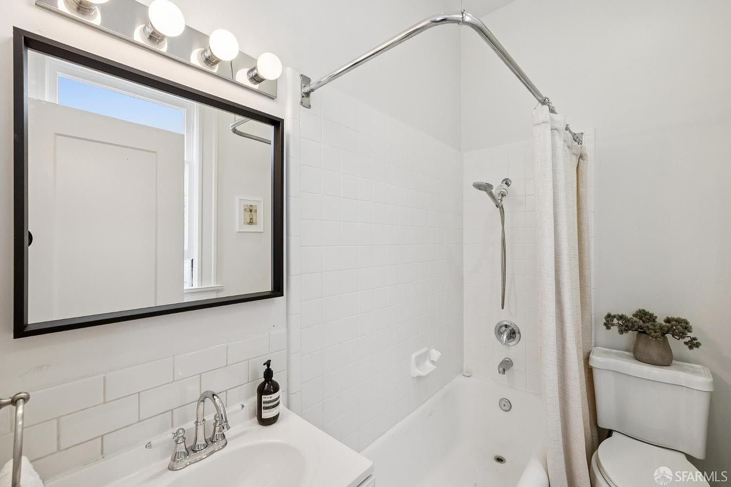 This is a bright and clean bathroom featuring white subway tile, a black-framed mirror with vanity lighting, and a white sink. The bathtub has a shower with a chrome showerhead and a neutral-colored shower curtain. A small plant sits on the toilet tank, adding a touch of nature to the space.