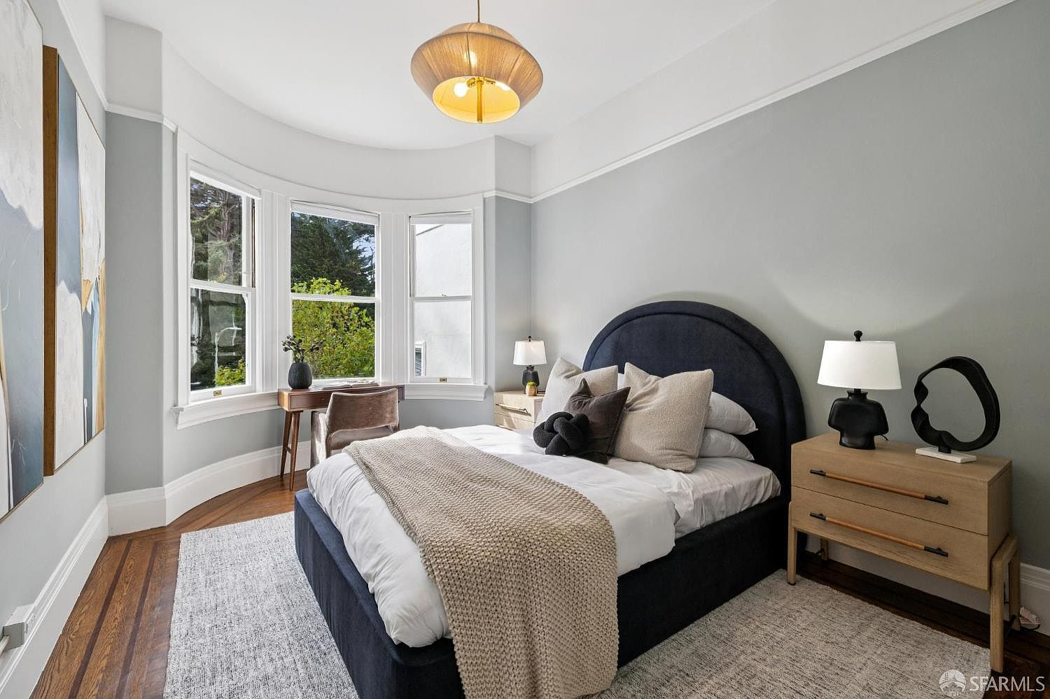This is a well-lit primary bedroom featuring a blue upholstered bed with multiple pillows and a beige knit throw. A desk sits by the bay window, and a wooden nightstand with a lamp and decorative object is positioned next to the bed. The room is decorated in neutral tones with hardwood floors and a light gray rug, creating a calm and inviting atmosphere.