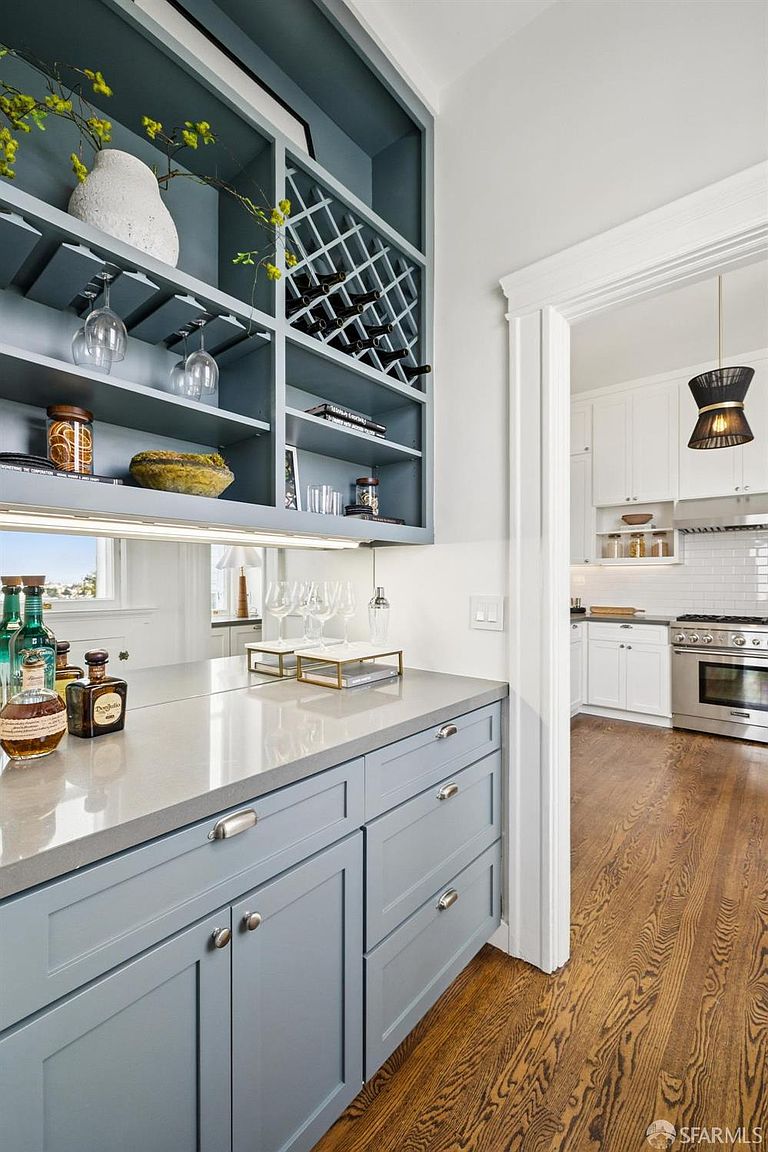 This interior shot showcases a stylish kitchen area with custom blue-grey cabinetry and a light grey countertop. The upper cabinets feature a built-in wine rack and glass storage, while the lower cabinets provide ample storage space. The kitchen opens into another area, revealing white cabinets and stainless steel appliances, creating a bright and inviting atmosphere.