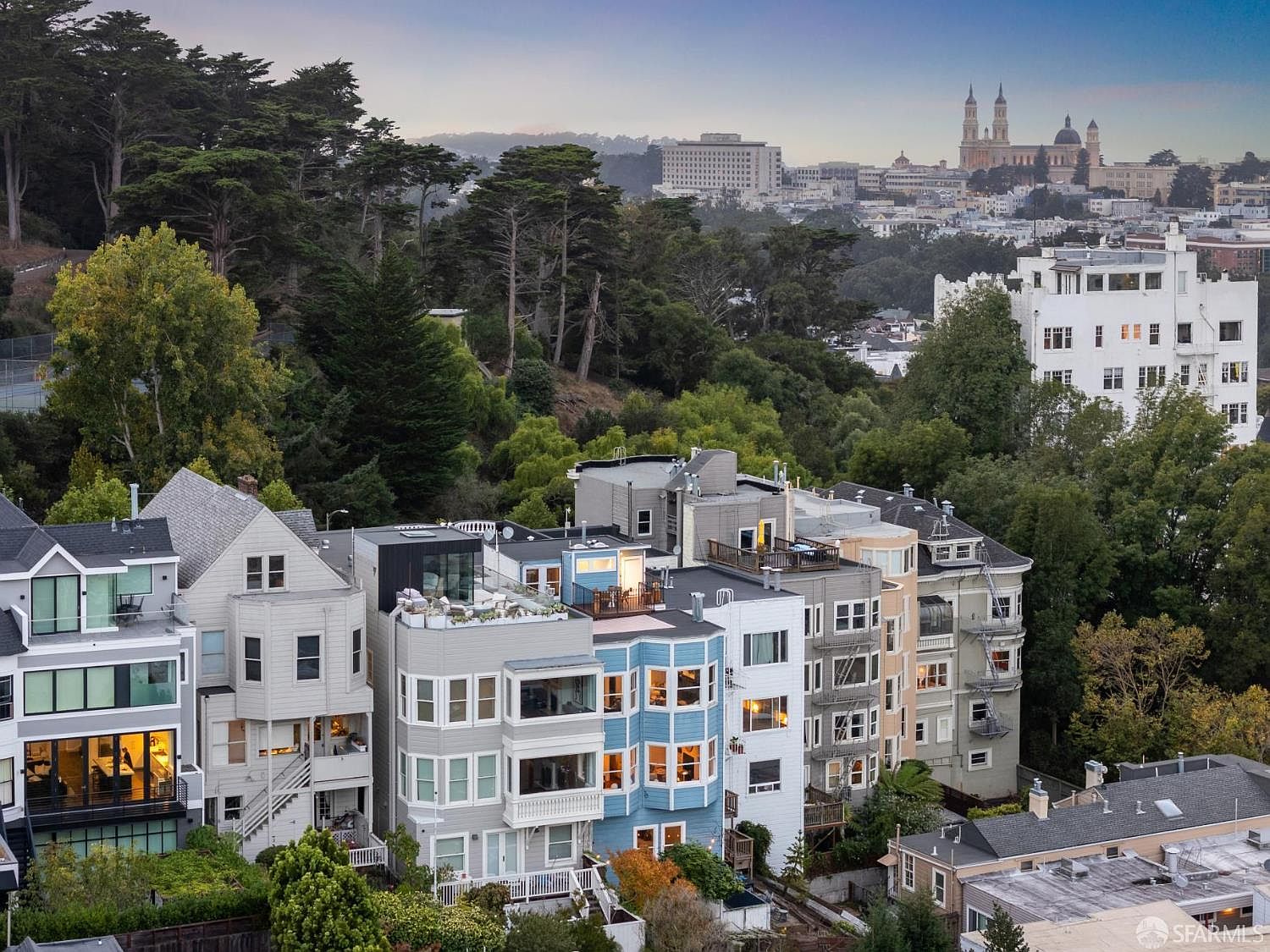 This aerial view showcases a row of colorful Victorian-style houses nestled among lush greenery in a hilly urban landscape. The houses feature bay windows, rooftop decks, and a variety of architectural details. In the background, a cityscape with prominent buildings and a church steeple adds depth and context to the scene.