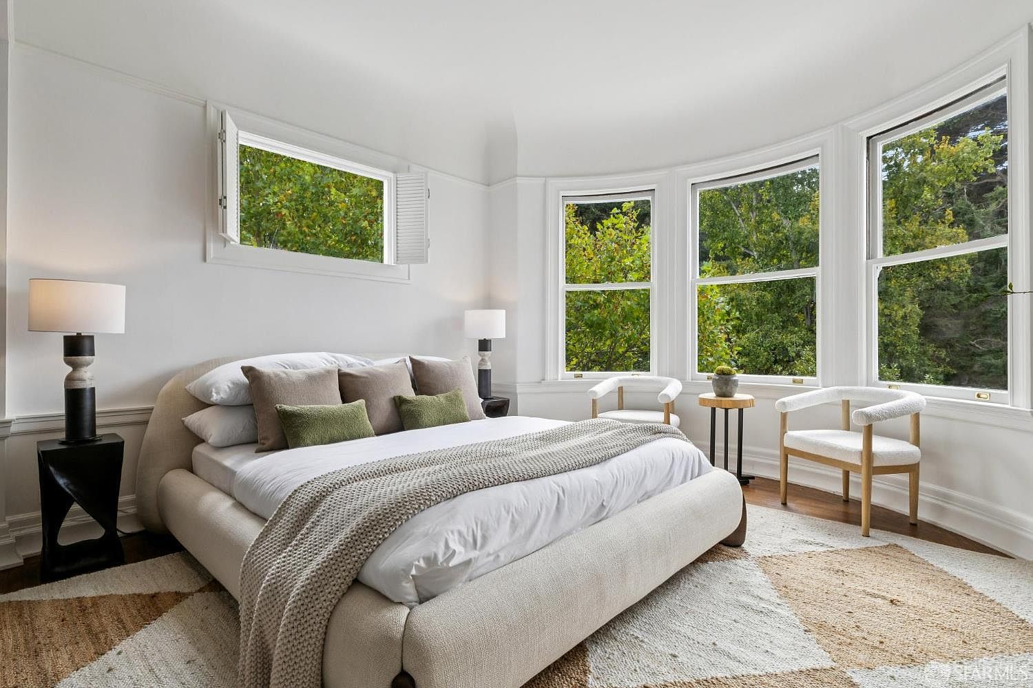 This is a bright and airy primary bedroom featuring a large bed with neutral-toned bedding and pillows. The room has multiple windows offering views of lush greenery, and the decor is minimalist and elegant. A patterned rug adds texture to the hardwood floors, creating a serene and inviting atmosphere.