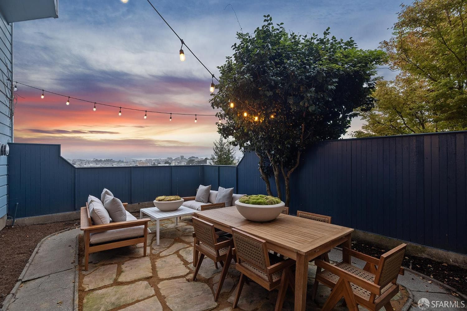 This image showcases a charming outdoor patio area, perfect for relaxation and entertaining. The space features a stone patio, comfortable seating arrangements with neutral cushions, and a wooden dining table set with chairs. String lights overhead add a warm, inviting ambiance, while a lush tree provides shade and privacy.