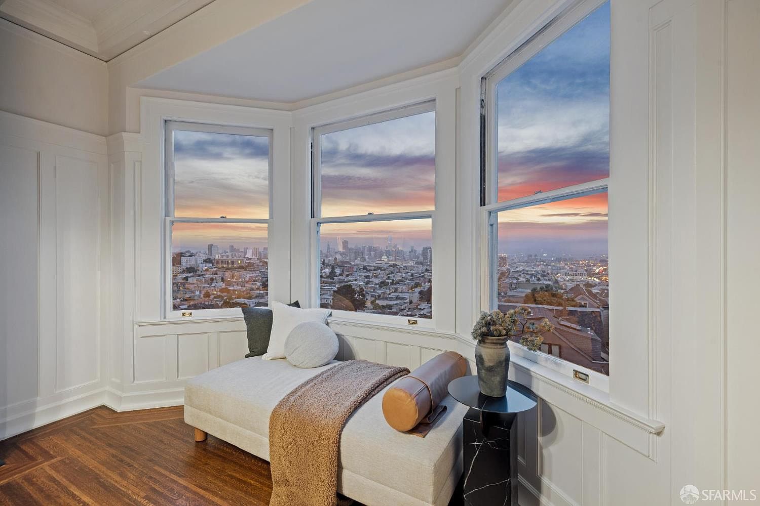 This interior shot showcases a cozy living space with a bay window offering a stunning city view at sunset. The room features a chaise lounge adorned with pillows and a throw, complemented by a small side table with a vase of flowers. The hardwood floors and paneled walls add a touch of classic elegance to the space.