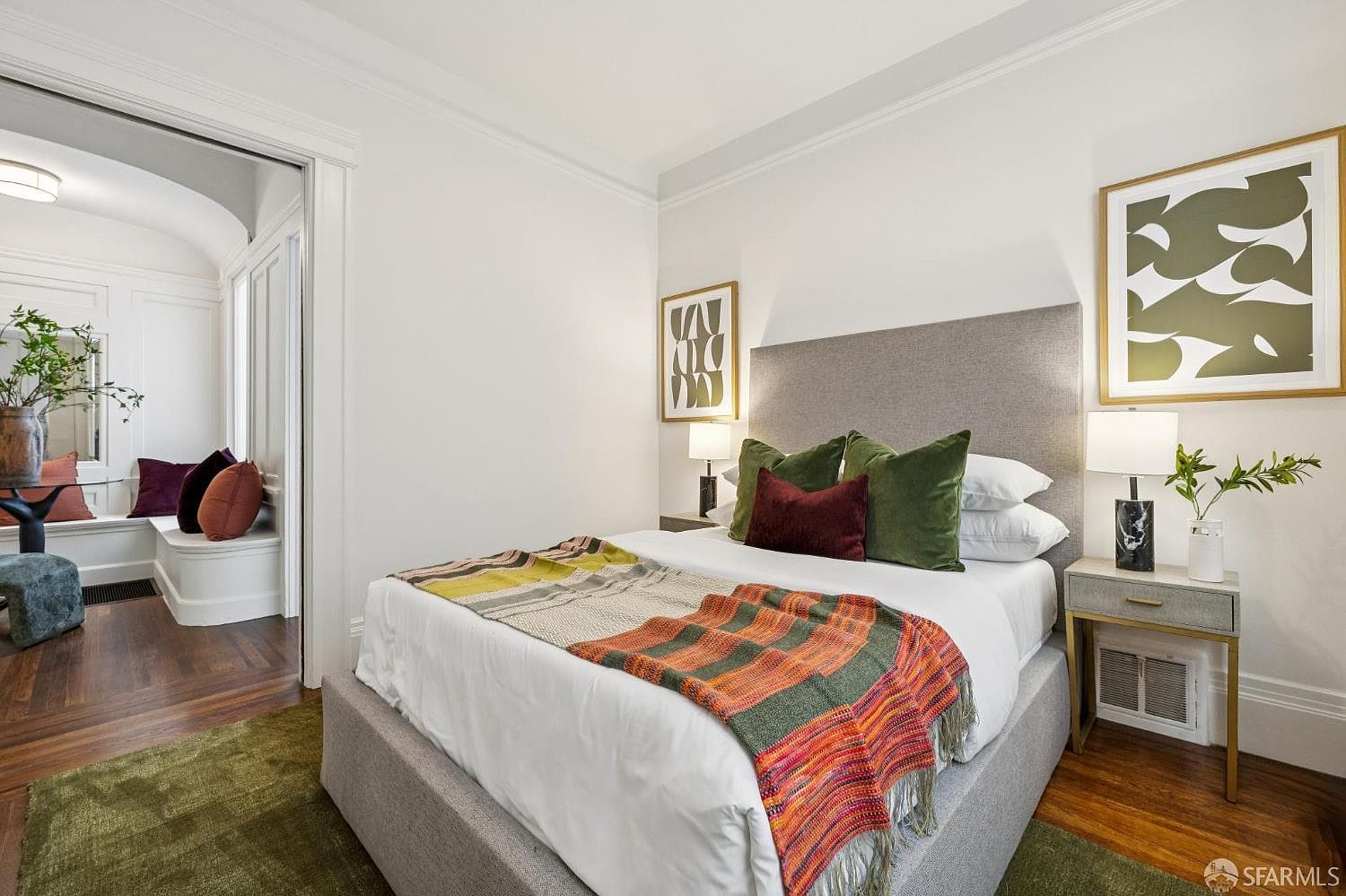 This is a cozy primary bedroom featuring a bed with a gray upholstered headboard, white linens, and a colorful throw blanket. The room is decorated with framed artwork and bedside tables with lamps. The hardwood floors and a glimpse into an adjacent sitting area add to the room's charm and appeal.