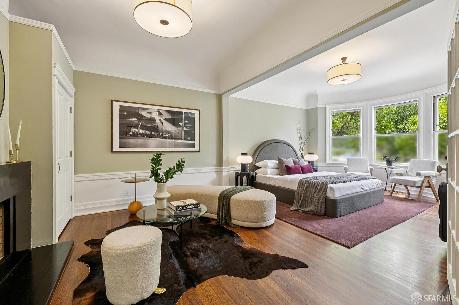 This is a spacious primary bedroom featuring a modern design with a calming color palette. The room is divided into a sleeping area and a sitting area, with a large bed, stylish seating, and ample natural light from the windows. The hardwood floors and area rug add warmth and texture to the space.