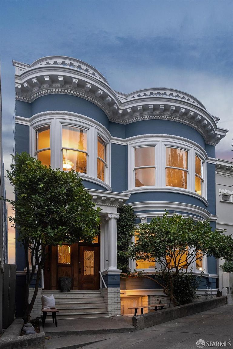 This is a captivating front view of a two-story Victorian-style house, showcasing its elegant blue facade, white trim, and curved bay windows. The entrance is framed by white columns and a wooden door, while mature trees add a touch of greenery and curb appeal. The overall impression is one of classic charm and sophistication.