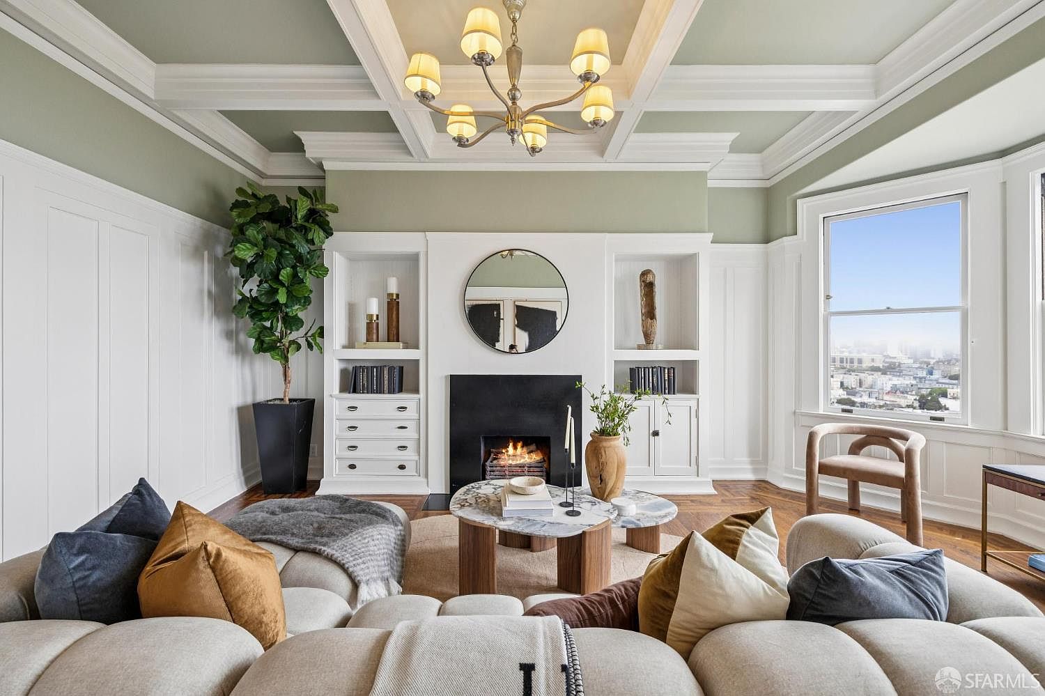 This is an interior shot of a living room featuring a coffered ceiling with a chandelier, a fireplace with built-in shelving, and a large sectional sofa. The room is decorated in neutral tones with pops of color from pillows and accessories, creating a cozy and inviting atmosphere. A large window provides natural light and a view of the city.