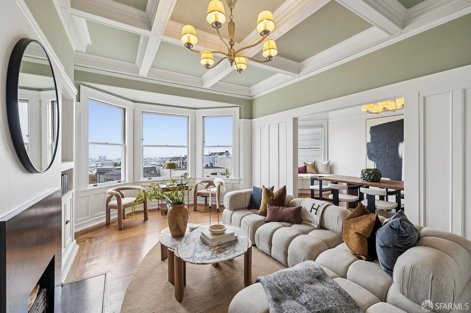 This is an interior shot of a well-lit living room featuring a coffered ceiling, a bay window with city views, and a modern, curved sofa. The room is decorated in neutral tones with pops of color from throw pillows and artwork, creating a sophisticated and inviting atmosphere. A round marble coffee table sits atop a large area rug, completing the stylish and comfortable living space.