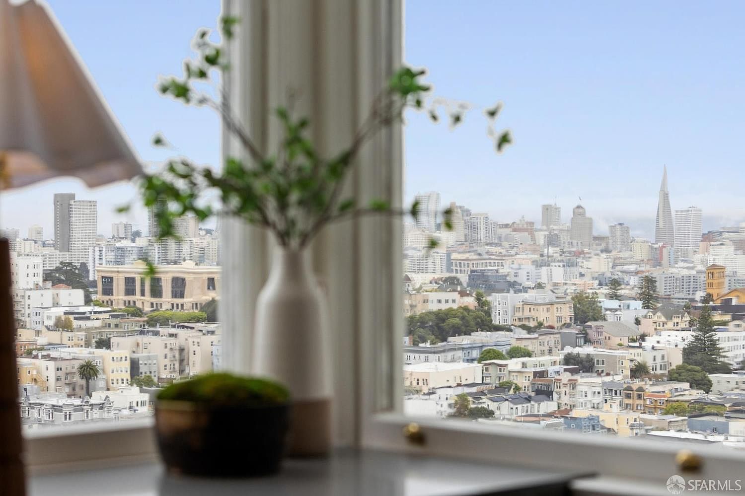 This image showcases a living room view through a window, overlooking a cityscape with numerous buildings and a clear sky. A decorative vase with greenery and a small potted plant are placed on a surface in the foreground, adding a touch of nature to the interior setting. The overall impression is one of urban elegance and a connection to the city.