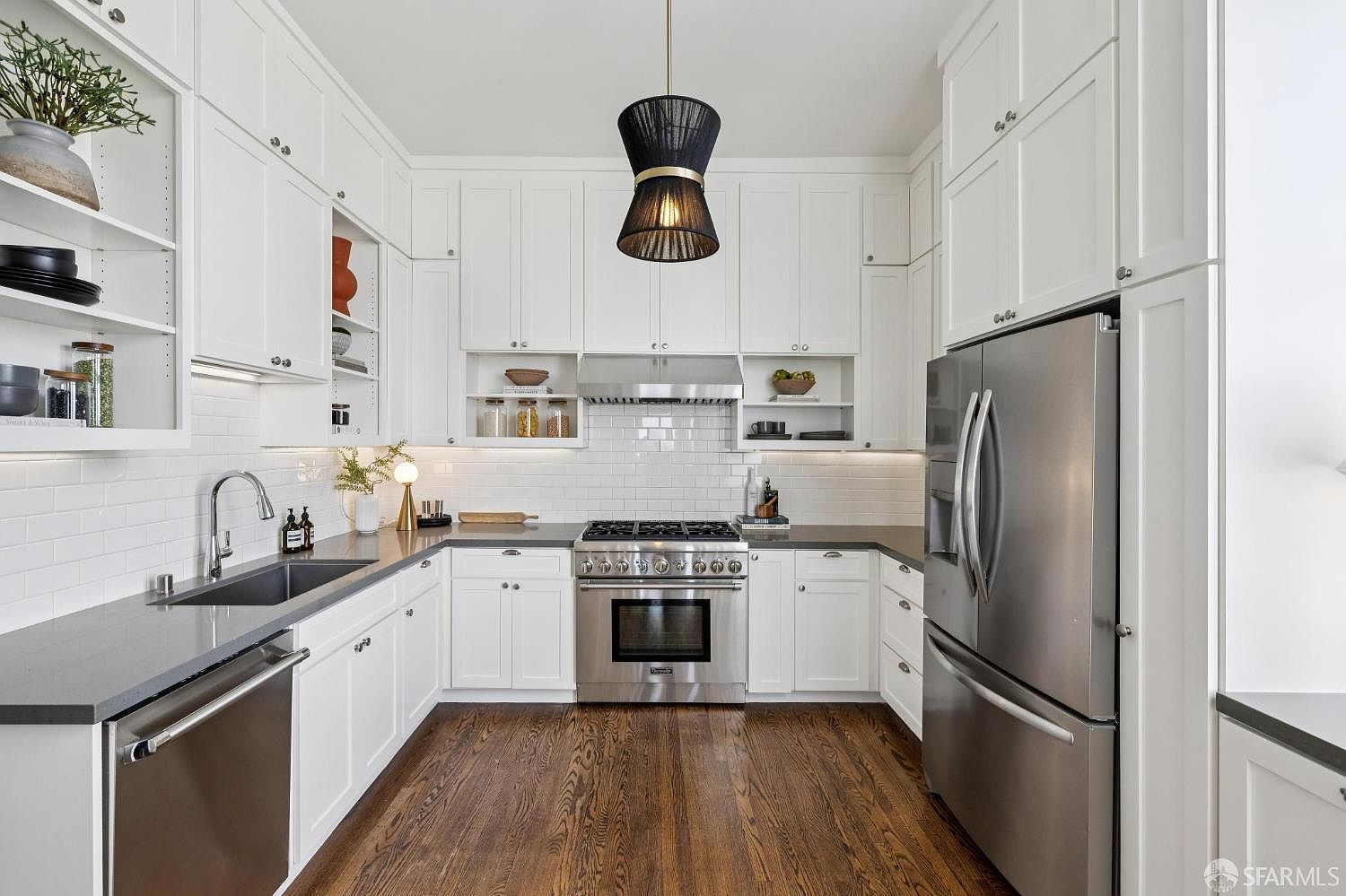 This is a well-lit kitchen featuring white cabinetry, stainless steel appliances, and dark countertops. The kitchen has a modern design with a black pendant light fixture hanging from the ceiling. The hardwood floors add warmth to the space, and the overall impression is clean and functional.