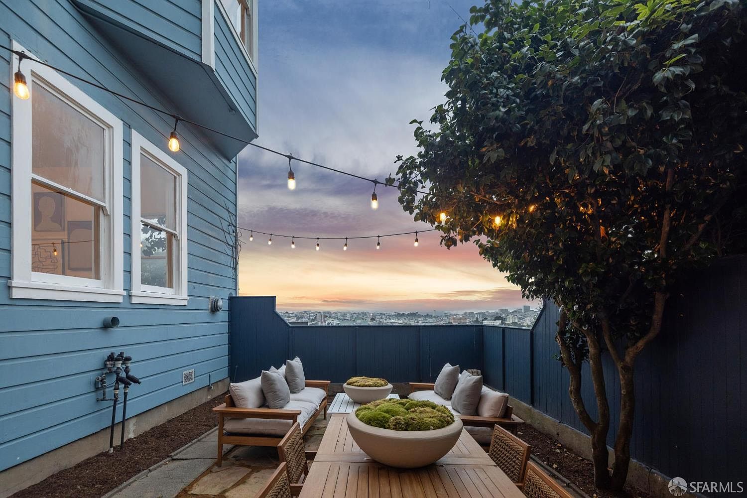 This image showcases a charming outdoor patio area, perfect for relaxation and entertaining. The space features comfortable seating arrangements with neutral-toned cushions, a wooden dining table adorned with decorative moss bowls, and string lights that add a warm ambiance. A lush tree provides shade and privacy, while the backdrop reveals a cityscape view at sunset, enhancing the property's appeal.