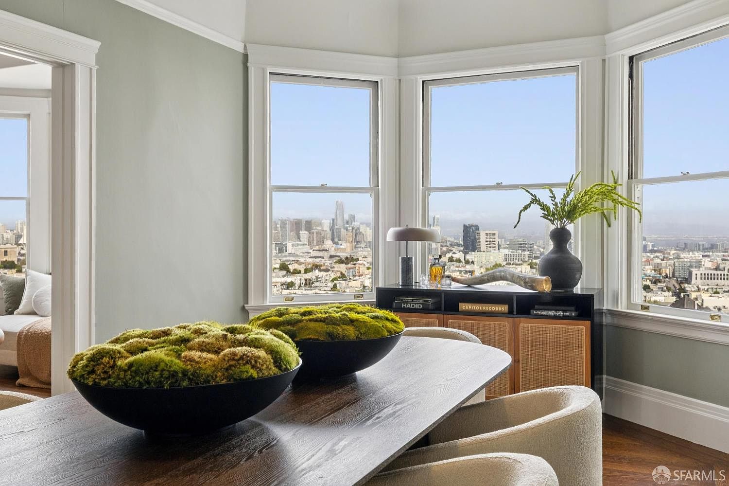 This image showcases a dining room with a modern aesthetic, featuring a dark wood table adorned with decorative moss bowls. Large windows offer a stunning city view, complemented by a stylish console table with a lamp and decorative items. The room is bathed in natural light, creating an inviting and sophisticated atmosphere.