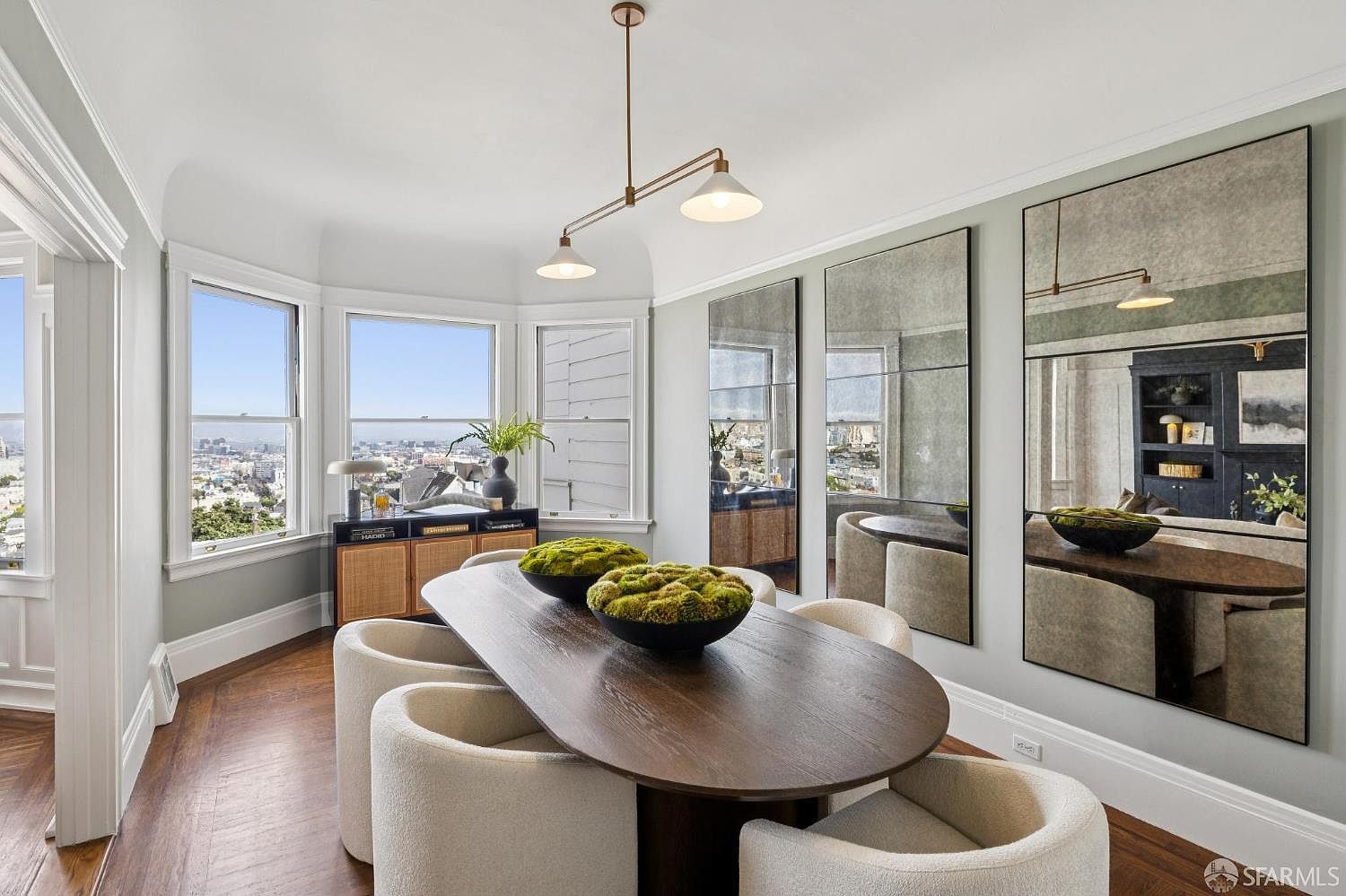 This is an interior shot of a dining room featuring a long, oval-shaped wooden table surrounded by plush, cream-colored chairs. The room is well-lit with natural light streaming through a bay window offering a city view, complemented by a modern pendant light fixture. Three large mirrors adorn the wall, reflecting the table and chairs, enhancing the sense of space and elegance.